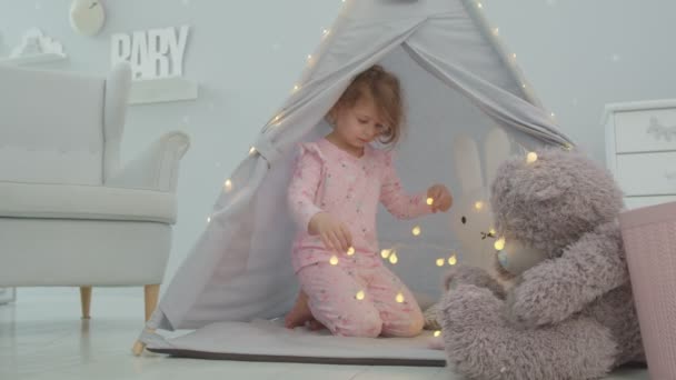 Νηπιοκόμος σε ροζ πιτζάμα παίζει με αρκουδάκι και φωτισμό γιρλάντα κάθεται σε παιδική σκηνή στο παιδικό δωμάτιο. Παιδικά παιχνίδια δαπέδου. — Αρχείο Βίντεο