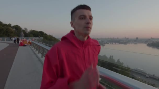 Millennial-Mann in roter Sportjacke läuft frühmorgens auf die Straße. Sonnenaufgang-Lauftraining. Gesunder Lebensstil. — Stockvideo