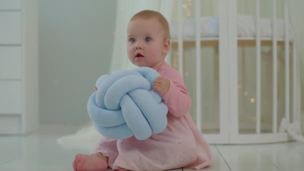 Spädbarn flicka i rosa klänning sitter och leker med mjuk leksak bollen på golvet i ljusa barn rum. Söt 10 månader gammal baby med stora blå ögon. — Stockvideo