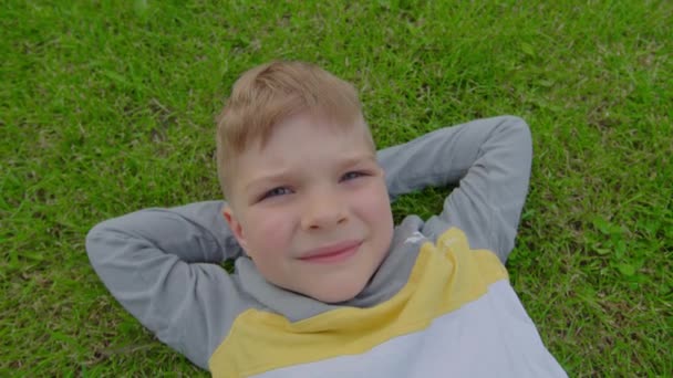 Ragazzo della scuola sdraiato sull'erba con la mano sopra la testa. Bambino di 6 anni che si rilassa a terra e guarda la fotocamera. — Video Stock