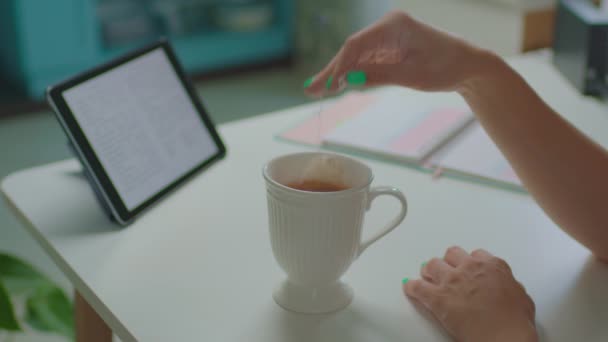 Té femenino de la mano mientras que lee el texto en la computadora de la tableta. Mujer sumergiendo la bolsa de té en una taza grande con agua caliente trabajando en la mesa. — Vídeos de Stock