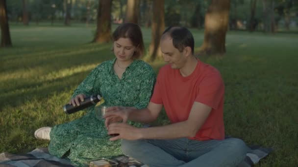 Vrouw in groene jurk gieten koffie in bekers zitten op picknick deken in het park. jong volwassen paar met picknick. — Stockvideo