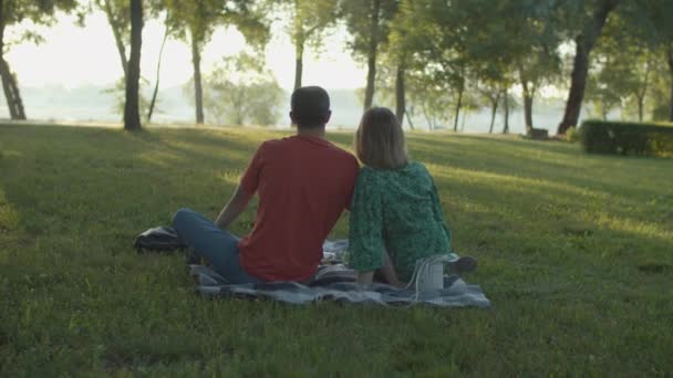 Visszapillantás a harmincas évekbeli párra, akik kora reggel piknikeznek a parkban. Steadicam lövés a család ül a takarón a parkban nézi a napfelkeltét. — Stock videók