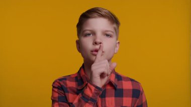 Genç bir çocuk sessiz kalmak için parmağını ağzının üzerine koyar. Parlak sarı arka plan onun ifadesini vurguluyor, neşeli ve eğlenceli bir ortam yaratıyor..