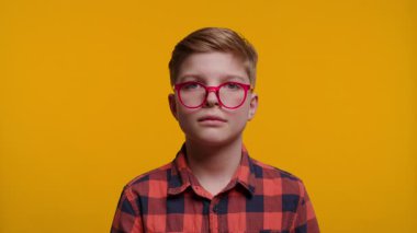 Neşeli çocuk gözlüğünü parmağıyla düzeltiyor ve parlak sarı bir arka planda kameraya gülümsüyor..