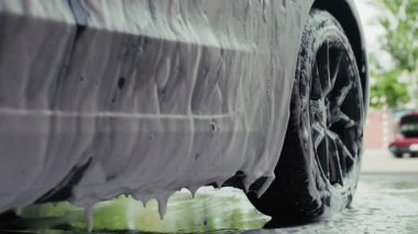 Araba yıkamada tamamen köpükle kaplanmış bir şekilde duruyor, kalın beyaz köpükler yavaşça yüzeye damlıyor, temizlik ve detaylara vurgu yapıyor..