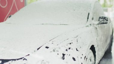 Bir araba, bir yıkama istasyonunda kalın beyaz köpükle kaplıdır. Araç bakımının temizlik ve detaylarını vurgular..