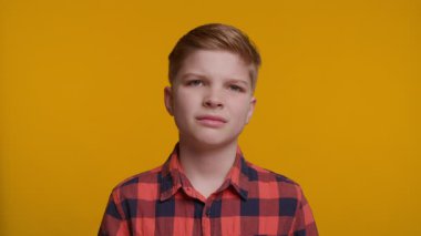 Çocuğun buruşuk bir yüzü var, rahatsızlık ya da iğrenme belirtileri gösteriyor. Arkaplan koyu sarı bir renktir. Kısa, düzgün taranmış saçları ve açık tenli genç bir çocuk kırmızı ve siyah bir ekose gömlek giyiyor..