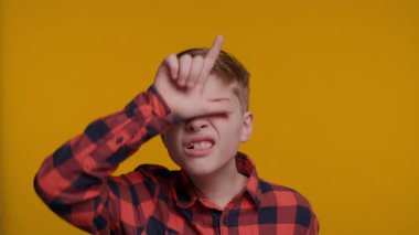 Genç bir çocuk L şeklini alıyor. Parmakları alnında ve muzip bir şekilde Ezik diyor. Onun ifadesi parlak sarı arka plana karşı kendinden emin ve haylaz.