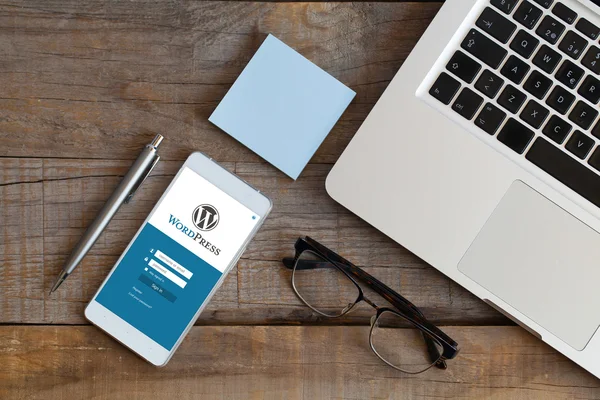 Wordpress app in uno schermo del telefono cellulare. Vista dall'alto della scrivania in legno con computer, occhiali, penna e posta . — Foto Stock