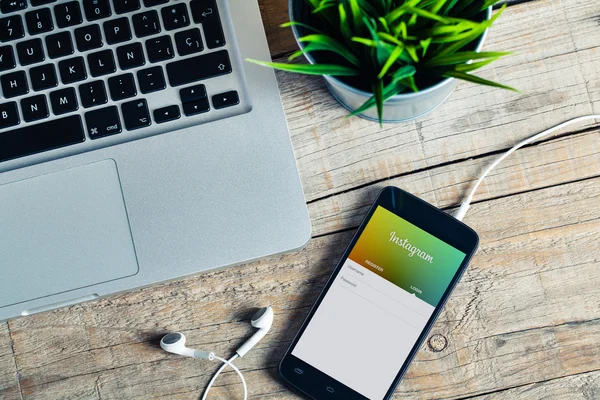 MALAGA, SPAIN - OCTOBER 29, 2015: Instagram app in a smart phone screen. Workplace with laptop earphones and a plant, over a wood table. — Stock Photo, Image