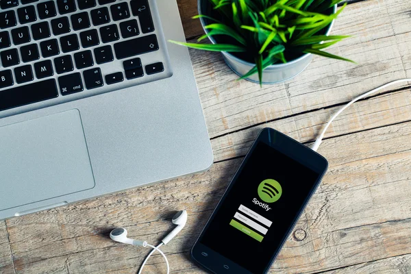 Malaga, Spanien - 29. Oktober 2015: Spotify-App auf dem Smartphone-Bildschirm. Arbeitsplatz mit Laptop-Kopfhörern und einer Pflanze, über einem Holztisch. — Stockfoto