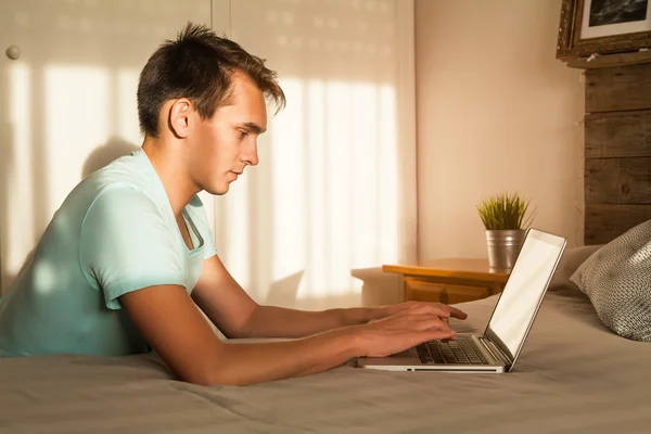 Joven hombre rubio escribiendo en un ordenador portátil en el dormitorio . —  Fotos de Stock
