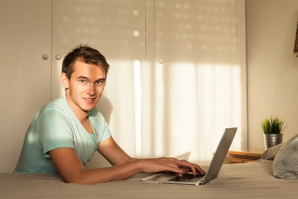 Junger blonder Mann tippt im Schlafzimmer in einen Laptop. — Stockfoto