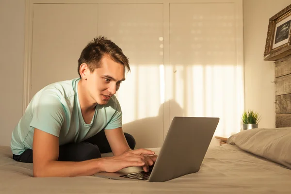 微笑的年轻金发碧眼的男人，在卧室里使用一台笔记本电脑. — 图库照片