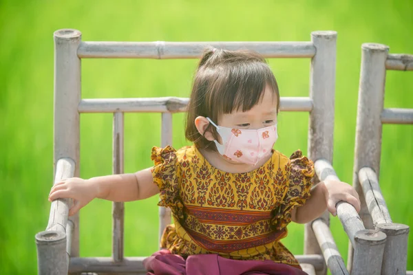 Little asian girl wear protect mask for children sitting on wood chair in green rice field