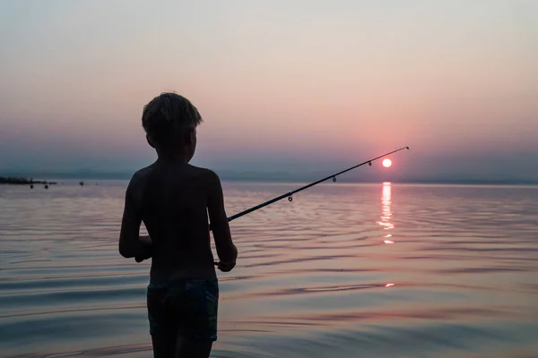 Ragazzo pesca con una canna da pesca in piedi in acqua la sera al tramonto Foto Stock Royalty Free