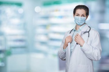 Yüzünde koruyucu bir maske olan doktor kadın bulanık bir arka planda duruyor..