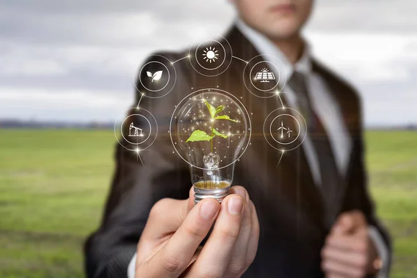 Businessman shows natural sources of energy and a burning light bulb with a sprout on a blurred background.