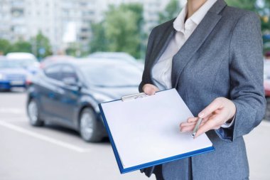 Bir iş adamı park halindeki arabaların yanında elinde bir not panosu ve kalemle duruyor..