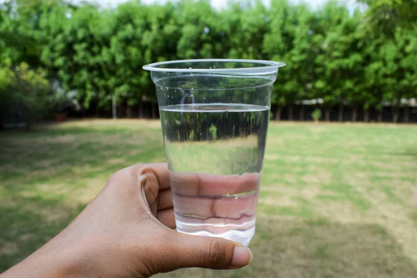 Female holding glass of clean filtered water in transparent disposable glass at home garden with background of Nature open environment of greenery lawn and trees. Save water,conserve water.safe nature