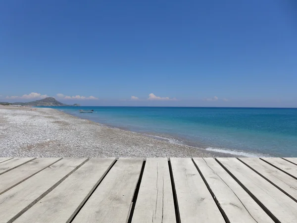 Ahşap iskele mavi deniz, gökyüzü ve beyaz çakıl plaj arka plan ile. Akdeniz manzara güneşli gün. Yunan Adası. — Stok fotoğraf