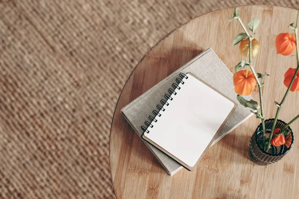 Empty diary, notepad mockup and book on wooden table. Cape gooseberry, dry physalis in vase. Blurred beige jute carpet background. Flat lay, top view. Simple Scandinavian boho interior.
