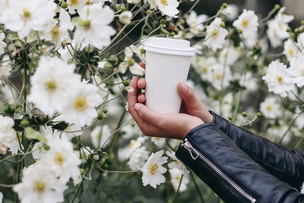 Weibliche Hände halten Kaffee zum Mitnehmen. Leere weiße Papierbecher-Attrappe mit Plastikdeckel. Nahaufnahme einer Frau in schwarzer Lederjacke inmitten blühender weißer Anemonenblumen. Selektiver Fokus. — Stockfoto
