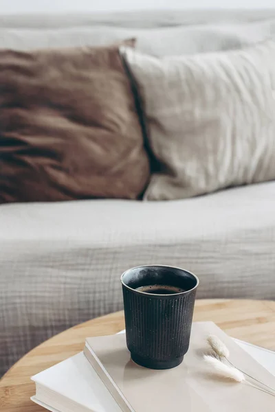 Books, dry bunny tail grass and black cup of coffee on round wooden table. Blurred beige sofa background with linen cushions. Hygge, cozy lifestyle concept. Scandinavian interior. Vertical. — Stock Photo, Image