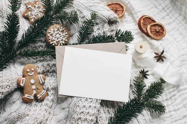 Christmas festive still life. Blank greeting card, invitation mockup and envelope on knitted sweater . Gingerbread cookies, dry oranges and fir branches on wool blanket. Selective focus, top view. — Stock Photo, Image