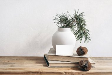 Noel hala hayattır. Eski ahşap bankta, masada kartvizit modeli. Çam ağacı dalları olan modern beyaz seramik vazo. Kahverengi Noel süsleri ve kitaplar. Beyaz duvar arkaplanı.
