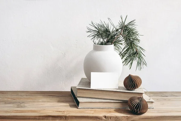 Christmas still life. Business card mockup on vintage wooden bench, table. Modern white ceramic vase with pine tree branches. Brown Christmas paper ornaments and books. White wall background. — Stock Photo, Image