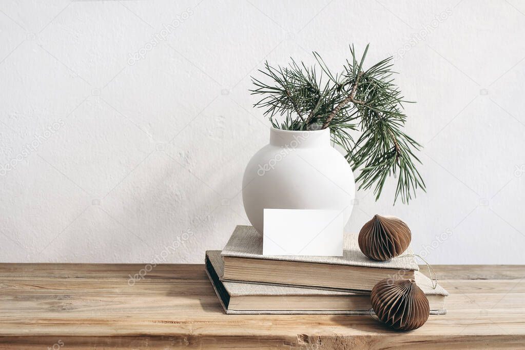 Christmas still life. Business card mockup on vintage wooden bench, table. Modern white ceramic vase with pine tree branches. Brown Christmas paper ornaments and books. White wall background.