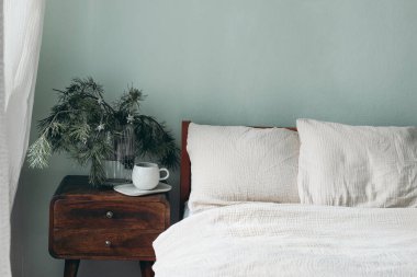 Christmas still life. Cup of coffee, retro wooden bedside table. Pine tree branches and silver stars ornaments in glass vase. Bed with beige cotton muslin beddings. Blank sage green wall background.