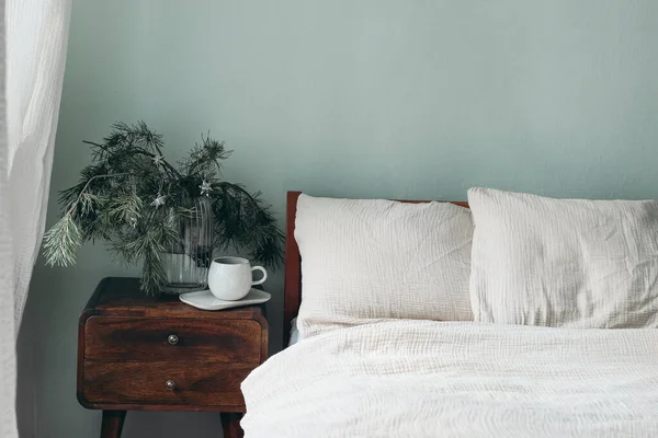 Natura morta di Natale. Tazza di caffè, comodino in legno retrò. Rami di pino e stelle d'argento ornamenti in vaso di vetro. Letto con lenzuola in mussola di cotone beige. Bianco verde salvia sfondo della parete. — Foto Stock