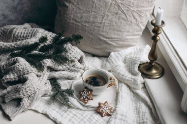 Sıcak kış kahvaltısı hayat sahnesi. Bir fincan sıcak kahve ve pencerenin yanında zencefilli kurabiye. Noel konsepti. Yastık, örgü süveter ve müslin ekose üzerinde köknar ağacı dalı. Klasik şamdan..