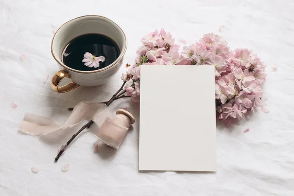 Wedding stationery mock-up. Blank greeting card and cup of coffee. White linen tablecloth background. Pink blossoming cherry tree branches and ribbon. Feminine still life composition. Flat lay,top. — Stock Photo, Image