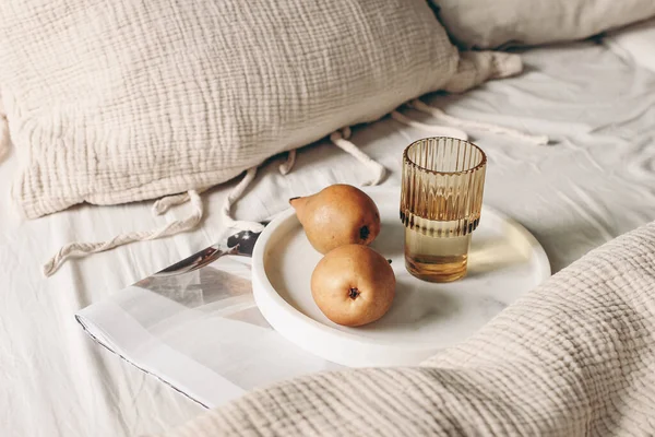 Autumn, summer breakfast in bed composition. Glass of water, folded newspapers and pear fruit on white marble tray. Champagne beige muslin cotton bed linen. Velvet cushions. Lifestyle, interior. — Stock Photo, Image