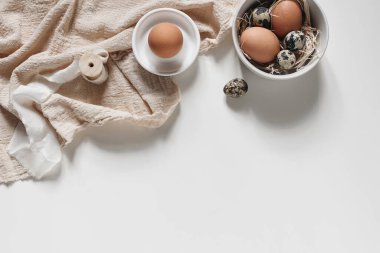Paskalya dekoratif çerçevesi, afiş. Bahar aylarında doğal bıldırcın yumurtaları, seramik kaseler ve beyaz masa arkasında izole edilmiş keten peçeteler. Tarafsız kadın stili fotoğraf, üst görünüm.