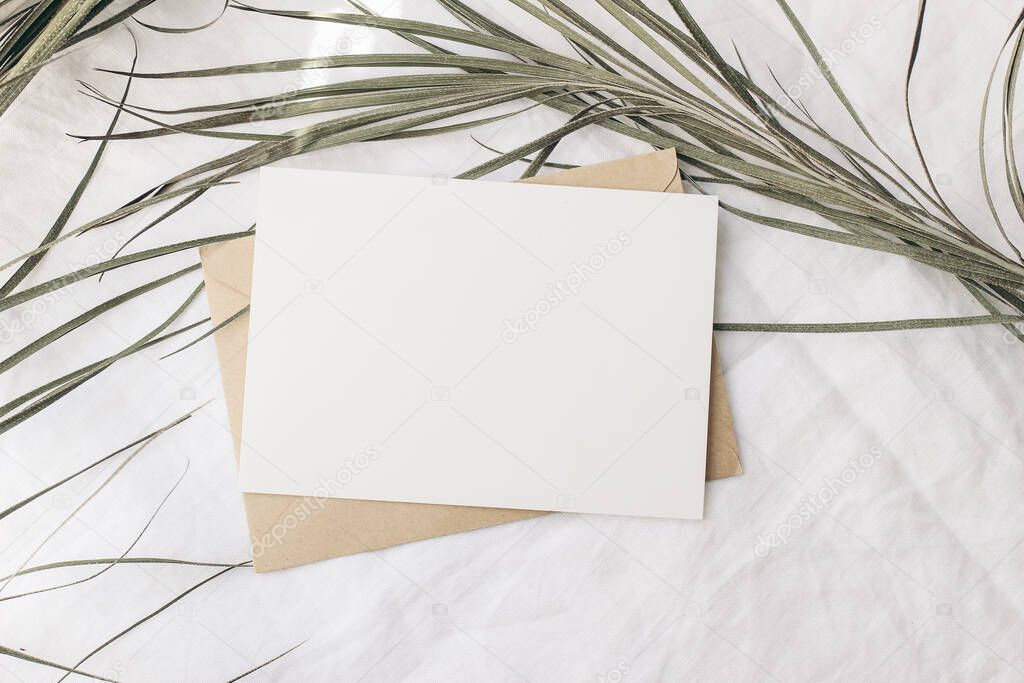 Summer tropical stationery still life scene. Dry date palm leaf on white table cloth in sunlight. Blank greeting card, invitation mockup with craft paper envelope. Long shadows. Vacation flat lay, top