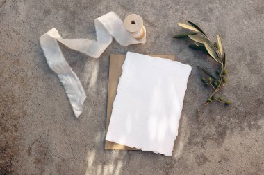 Yaz düğün kırtasiyesi, doğum günü modeli sahnesi. Boş tebrik kartı, beyaz ipek kurdele ve zeytin dalı. Güneş ışığında çamurlu beton arka plan, uzun gölgeler. Düz yatış, üst görünüm.