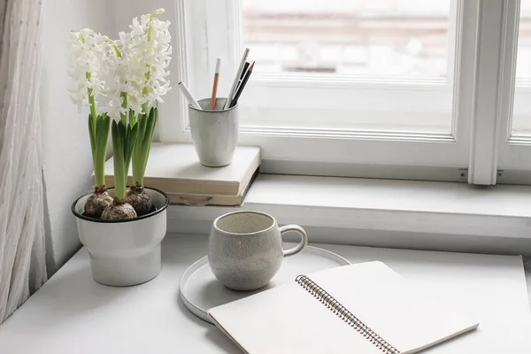 Easter spring still life. Cup of coffee, book and blank open diary mockup near window sill. White hyacinth in flower pot. Pencils in ceramic holder. Home office concept. Scandinavian interior