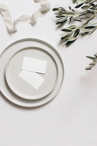 Summer wedding stationery still life scene. Blank business cards mock-ups on ceramic plates. Silk ribbon, olive tree branches isolated on white table background. Flat lay, top view. Empty copy space.