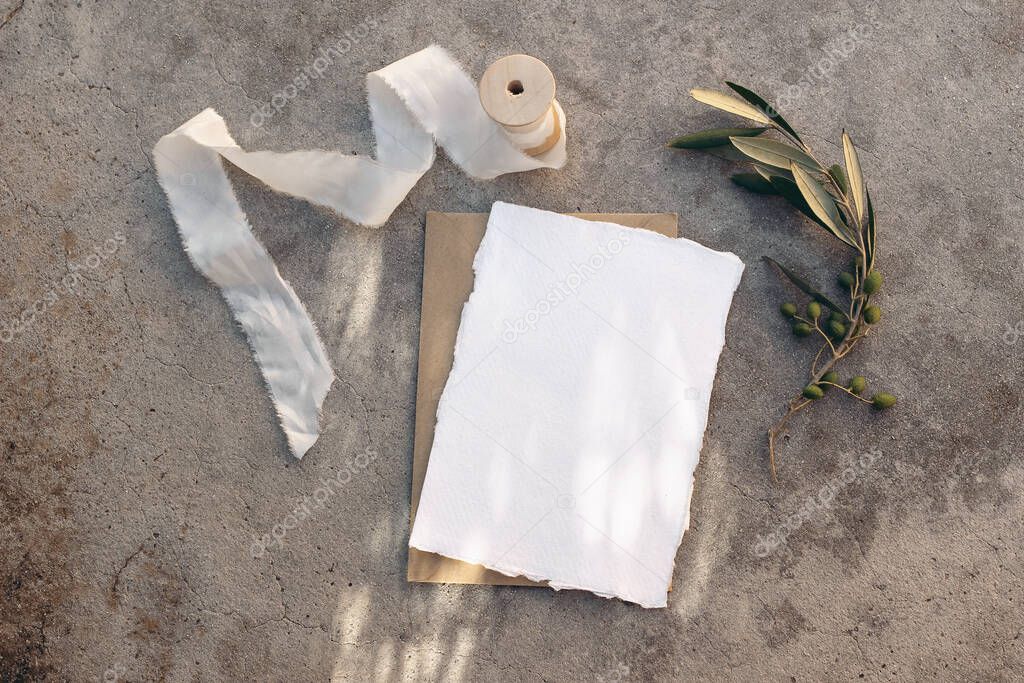 Summer wedding stationery, birthday mock-up scene. Blank greeting card, white silk ribbon and olive tree branches. Grunge concrete background in sunlight, long shadows. Flat lay, top view.