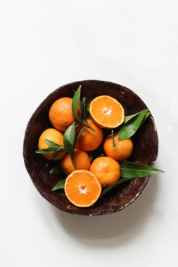 Taze portakal, mandalina. Kahverengi el yapımı seramik kasede yeşil yapraklı meyveler beyaz masa arkası. Sağlıklı çiğ gıda iskeleti, web afişi. Yaz kavramı. Dikey düzlük, üst