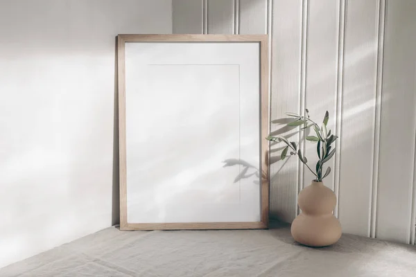 Portrait empty wooden frame mockup in sunlight. Olive branch in modern organic shaped vase. Beige linen table cloth. White wainscot wall paneling background. Scandinavian interior, home design. Art — Stock Photo, Image