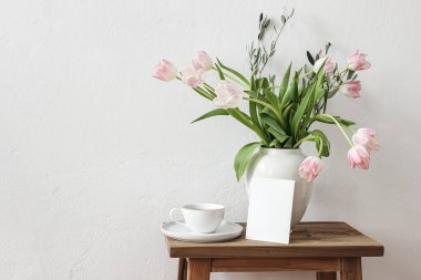 Kırsal Paskalya 'da hayat sahnesi. Boş tebrik kartı maketi, bir fincan kahve ve tahta bankta duran beyaz seramik vazoda çiçek buketi. Bahar fotoğrafı. Pembe laleler çiçekler, masada zeytin dalları..