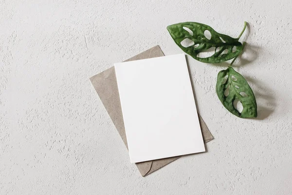 Summer wedding stationery mock-up scene. Blank greeting card, invitation with craft envelope and two little green monstera plant leaves. White textured background. Flat lay, top view. Minimal design.