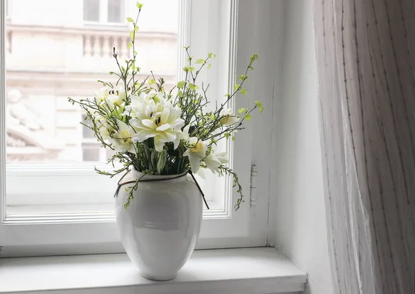 Easter spring still life. Floral bouquet on window sill. White and yellow tulips, daffodils flowers and green birch tree and blueberry branches in ceramic vase pot. Home decor. Scandinavian interior — Stock Photo, Image