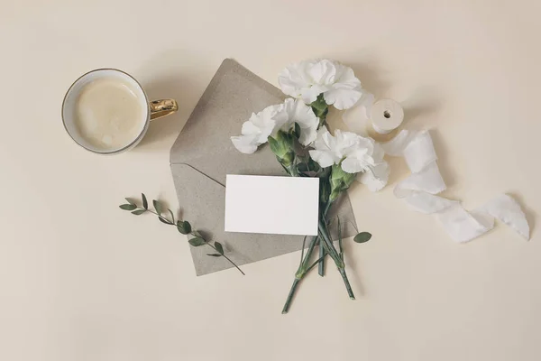 Composição feminina do café da manhã. Mockup de cartão de visita. Papelaria de casamento ou aniversário. Xícara de café, flores de cravo branco e ramos de eucalipto. Fundo da mesa bege. Deitado plano, vista superior. — Fotografia de Stock